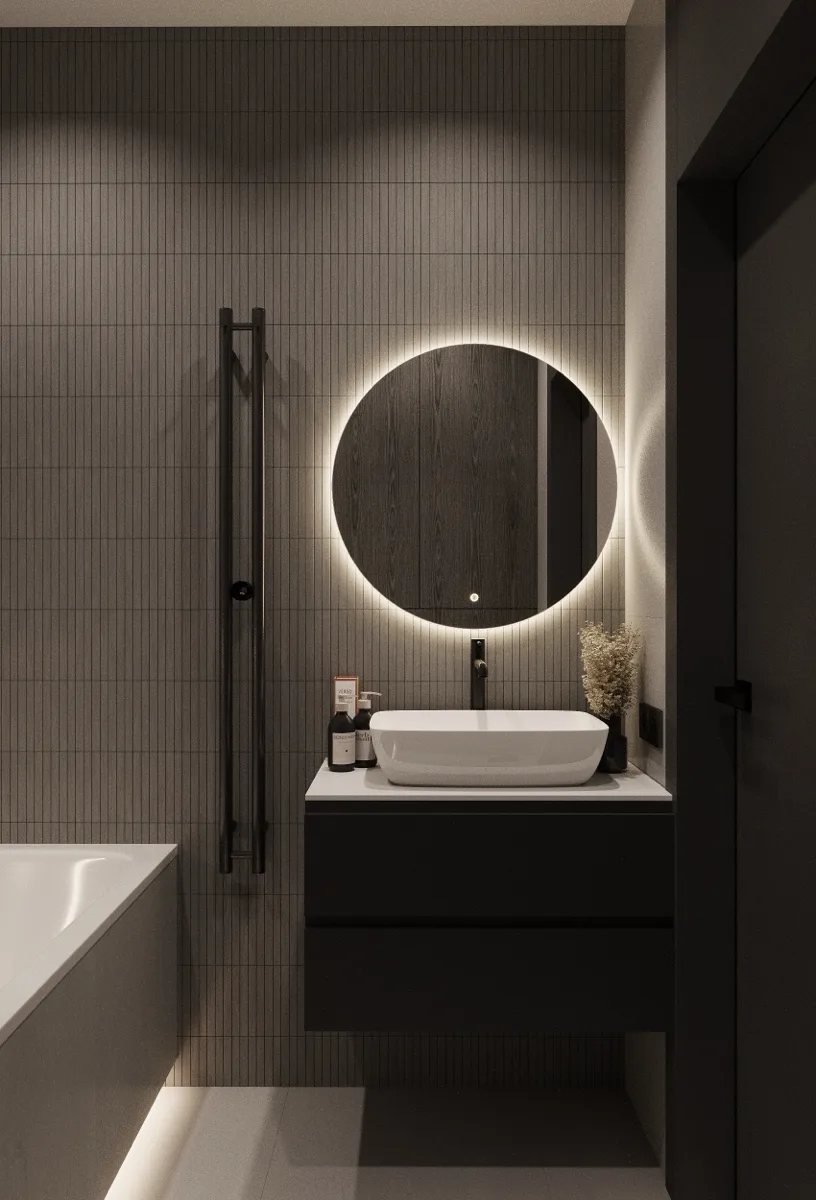 Санузел: серая плитка узкой вертикальной раскладкой, круглое зеркало с подсветкой, подвесная тумба с накладной раковиной, чёрный полотенцесушитель, ванна